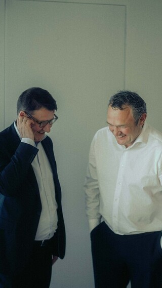 Zwei Personen in Business-Kleidung stehen in einem modernen Bürobereich und lachen miteinander.