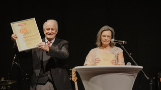 Ein Mann und eine Frau stehen auf der Bühne. Die Frau steht an einem Pult mit Mikrofon und hält Moderationskarten in der Hand, sie blickt in die Moderationskarten. Der Mann lächelt und hält freudig eine Auszeichnung in den Händen.