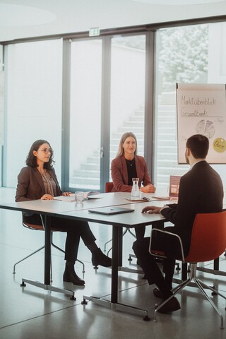 In einem Besprechungszimmer sitzen ein Mann und zwei Frauen.