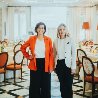 Zwei Damen in eleganter Kleidung stehen lächelnd in einem edlen Speisesaal.