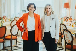 Zwei Damen in eleganter Kleidung stehen lächelnd in einem edlen Speisesaal.