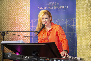 Sängerin spielt am Piano und singt. Sie trägt ein orangenes Kleid und hat blonde lange Haare.