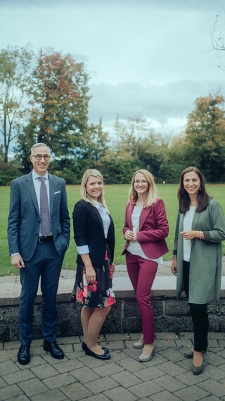 Vier Personen lächeln draußen in die Kamera, mit einer Wiese und Bäumen im Hintergrund.