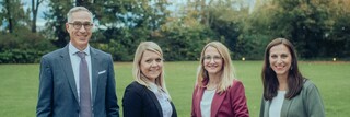 Vier Personen lächeln draußen in die Kamera, mit einer Wiese und Bäumen im Hintergrund.
