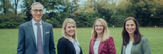 Vier Personen lächeln draußen in die Kamera, mit einer Wiese und Bäumen im Hintergrund.
