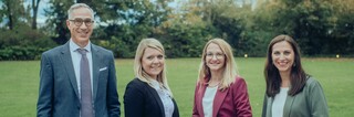 Vier Personen lächeln draußen in die Kamera, mit einer Wiese und Bäumen im Hintergrund.