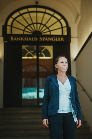 Eine Dame in Geschäftskleidung steht auf der Treppe vor einem metallenen Portal mit der Aufschrift "Bankhaus Spängler".