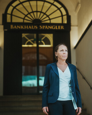 Eine Dame in Geschäftskleidung steht auf der Treppe vor einem metallenen Portal mit der Aufschrift "Bankhaus Spängler".