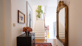Ein eingerichtetes Wohnhaus mit einer Treppe ins obere Stockwerk, einem großen goldenen Spiegel auf der rechten Seite sowie zwei Bildern und einer kleinen Holzkommode auf der linken Seite.