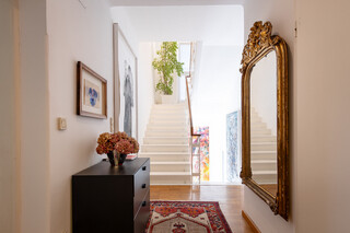 Ein eingerichtetes Wohnhaus mit einer Treppe ins obere Stockwerk, einem großen goldenen Spiegel auf der rechten Seite sowie zwei Bildern und einer kleinen Holzkommode auf der linken Seite.