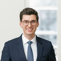 Ein lächelnder Herr in Anzug steht vor einem leeren Besprechungstisch.