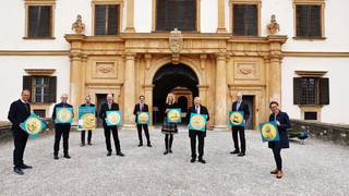 Mehrere Personen (7 Männer, 2 Frauen), alle dunkel gekleidet, stehen im Außenbereich vor einem historischen Gebäude (weiß mit beigem Stuck). Sie alle haben eine Auszeichnung in der Hand, auf welcher eine goldene Münze mit der Aufschrift "Das Gold der Erzbischöfe) in den Händen.