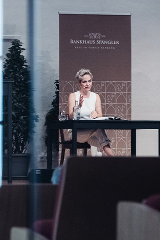 junge Frau im weißem Kleid sitzt auf einer Bühne und liest aus einem Buch vor.