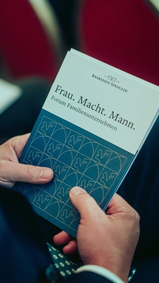 Die Hände eines Mannes sind sichtbar, während er eine Broschüre mit dem Text "Frau. Macht. Mann." beim Forum Familienunternehmen 2024 hält.