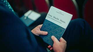 Die Hände eines Mannes sind sichtbar, während er eine Broschüre mit dem Text "Frau. Macht. Mann." beim Forum Familienunternehmen 2024 hält.