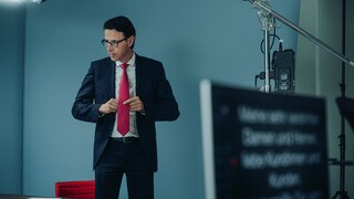 Dr. Nils Kottke steht im Anzug mit roter Krawatte vor einem Tisch, umgeben von Licht und Aufnahmegeräten