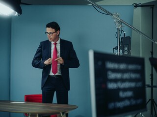 Dr. Nils Kottke steht im Anzug mit roter Krawatte vor einem Tisch, umgeben von Licht und Aufnahmegeräten