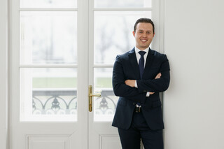 Ein lächelnder Herr in Anzug steht mit verschränkten Armen vor einer Balkontür.