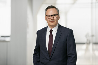 In einem hellen Raum steht ein Mann mit Brille und blauem Anzug.