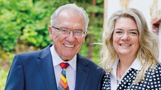 Älterer Mann mit grauem Harr und Brille, der freundlich lächelt. Er trägt ein dunkelgraues Sakko mit weißem Hemd und bunter Krawatte. Neben ihm steht eine Frau mit blonden, langen gewellten Haaren und Brille. Sie trägt eine schwarze Hose mit weißer Blus und einem schwarz-weißem Blazer. Im Hintergrund sind grüne Bäume ersichtlich.