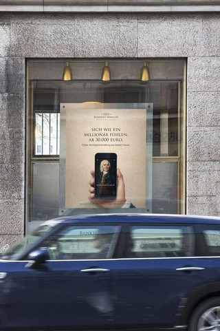 Dunkelblaues Auto (Marke: Mini) fährt an einem Gebäude des Bankhaus Spängler vorbei. Auslagefenster und ein Bankomat-Hinweisschild sind ersichtlich.