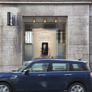 Dunkelblaues Auto (Marke: Mini) fährt an einem Gebäude des Bankhaus Spängler vorbei. Auslagefenster und ein Bankomat-Hinweisschild sind ersichtlich.