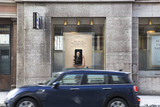Dunkelblaues Auto (Marke: Mini) fährt an einem Gebäude des Bankhaus Spängler vorbei. Auslagefenster und ein Bankomat-Hinweisschild sind ersichtlich.