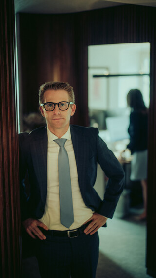 Ein Mann in seinen Dreißigern trägt ein weißes Hemd, eine hellblaue Krawatte und einen dunklen Anzug; er steht in einer Tür mit beiden Händen in die Hüften gestützt. Im verschwommenen Hintergrund steht eine Frau.