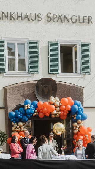 Elegant gekleidete Personen vor einem mit Luftballons geschmückten Portal eines historischen Gebäudes mit der Aufschrift "Bankhaus Spängler".