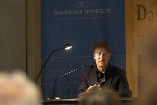 Tobias Moretti vor Hintergrund mit Aufschrift "Bankhaus Spängler". Er liest und schaut ins Publikum.