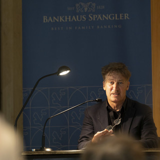 Tobias Moretti vor Hintergrund mit Aufschrift "Bankhaus Spängler". Er liest und schaut ins Publikum.