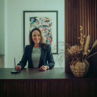 Eine Frau mittleren Alters mit dunklen schulterlangen Haaren, hellblauem Oberteil, dunklem Blazer und Brille steht hinter einem Pult; darauf liegen ein Kugelschreiber und eine Vase, im Hintergrund hängt ein Bild.