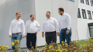 Vier Unternehmer im Freien vor einem Unternehmensgebäude, die sich gegenseitig ansehen. Eine Blumenwiese ist im Vordergrund ersichtlich.