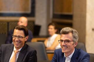 Zwei Herren in Anzug sitzen lachend auf Ledersesseln vor einer marmornen Wand.