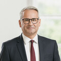 Mann mit grauem Haar und Brille lächelt im Stehen. Er trägt ein dunkelblaues Sakko mit weißem Hemd und einer weinroten Krawatte mit dunkelblauen Punkten.
