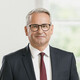 Mann mit grauem Haar und Brille lächelt im Stehen. Er trägt ein dunkelblaues Sakko mit weißem Hemd und einer weinroten Krawatte mit dunkelblauen Punkten.