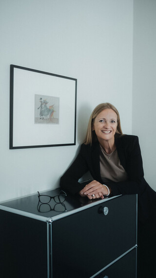 Person in Business-Kleidung lehnt an einer schwarzen Kommode und blickt in die Kamera.