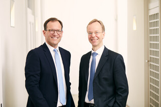 Zwei Männer stehen in einem hellen Büro. Beide tragen einen schwarzen Anzug mit weißem Hemd. Ein Mann hat dunkle Haare und trägt eine hellblaue Krawatte, der andere hat helle Haare und trägt eine dunkelblaue Krawatte. Beide tragen eine Brille und lächeln freundlich.