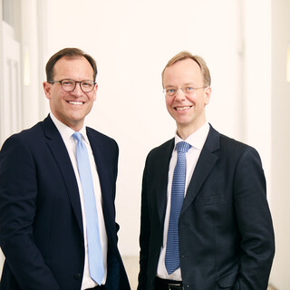 Zwei Männer stehen in einem hellen Büro. Beide tragen einen schwarzen Anzug mit weißem Hemd. Ein Mann hat dunkle Haare und trägt eine hellblaue Krawatte, der andere hat helle Haare und trägt eine dunkelblaue Krawatte. Beide tragen eine Brille und lächeln freundlich.