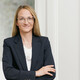 Frau mit blonden kürzeren Haaren lächelt und blättert in einer Broschüre. Sie trägt eine Brille, weiße Armbänder und ein dunkles Kleid.