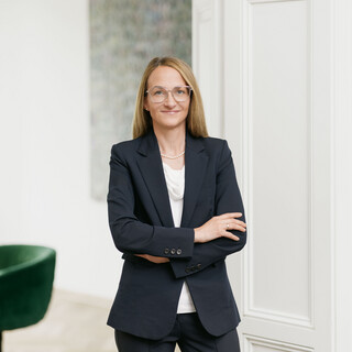 Frau mit blonden kürzeren Haaren lächelt und blättert in einer Broschüre. Sie trägt eine Brille, weiße Armbänder und ein dunkles Kleid.