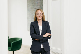 Frau mit blonden kürzeren Haaren lächelt und blättert in einer Broschüre. Sie trägt eine Brille, weiße Armbänder und ein dunkles Kleid.