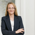 Frau mit blonden kürzeren Haaren lächelt und blättert in einer Broschüre. Sie trägt eine Brille, weiße Armbänder und ein dunkles Kleid.