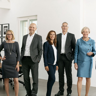 Mehrere Personen (5 Frauen, 3 Männer) stehen in einem lichtdurchfluteten Besprechungssaal. Im Hintergrund ist ein großer Tisch mit mehreren Bürostühlen.
