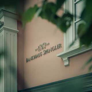 Detailaufnahme der Fassade des Bankhaus Spängler: Durch die Blätter hindurch ist an der rosafarbenen Fassade der Schriftzug Bankhaus Spängler zu sehen.