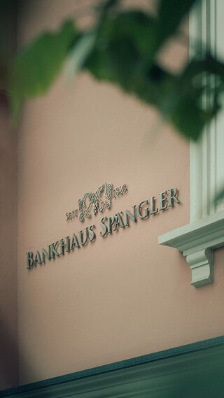 Detailaufnahme der Fassade des Bankhaus Spängler: Durch die Blätter hindurch ist an der rosafarbenen Fassade der Schriftzug Bankhaus Spängler zu sehen.