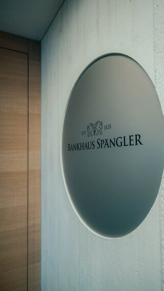 Runde Metallscheibe mit dem Logo und Schriftzug „Bankhaus Spängler“ an einer hellen Wand.