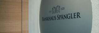 Runde Metallscheibe mit dem Logo und Schriftzug „Bankhaus Spängler“ an einer hellen Wand.