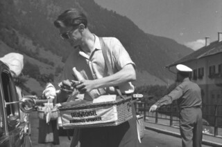 Ein historisches Schwarz-Weiß-Foto zeigt einen sommerlich bekleideten Herrn mit einem umgehängten Korb mit dem er auf einer Straße Geldwechsel anbietet.