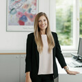 Freundlich lächelnde Frau mit langen brünetten Haaren steht vor einem Schreibtisch. Sie trägt einen schwarzen Hosenanzug und eine weiße Bluse. Im Hintergrund befindet sich ein farbiges Bild und ein grauer Büroschrank sowie ein PC am Schreibtisch.